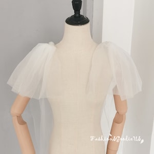 May include: White tulle bridal bolero with puffed sleeves. The bolero is on a white dress form.