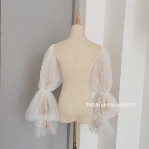 May include: White tulle bridal sleeves with puffed, gathered, and ruffled details. The sleeves are long and have a sheer, delicate look.