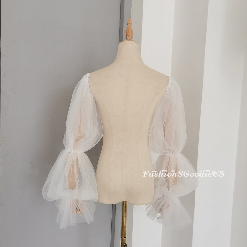 May include: White tulle bridal sleeves with puffed, gathered, and ruffled details. The sleeves are long and have a sheer, delicate look.
