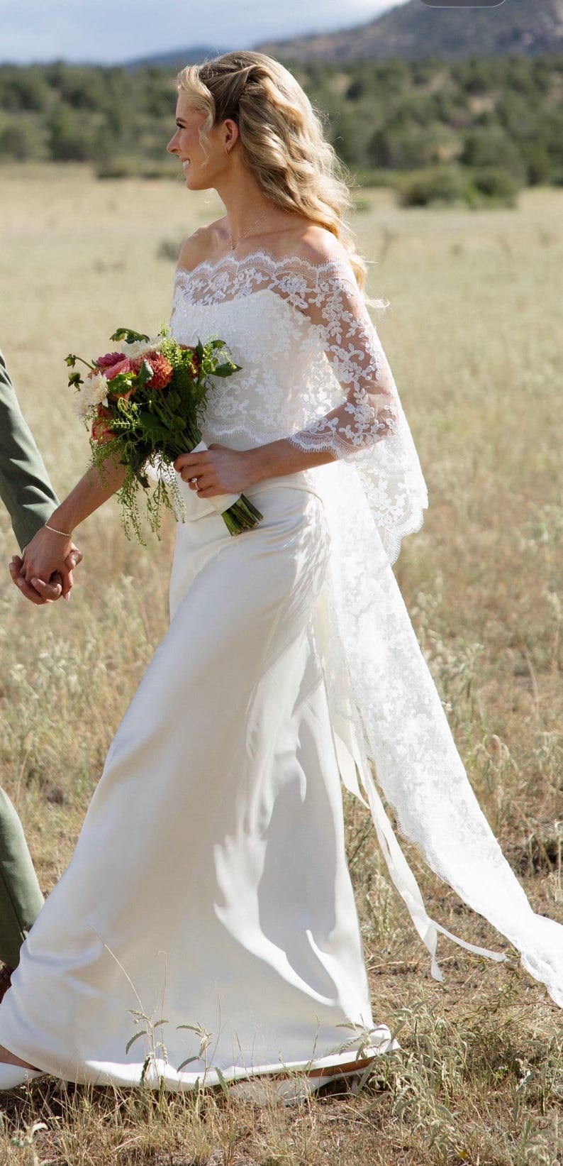 May include: A white floor-length gown featuring an off-the-shoulder neckline and long sleeves crafted from intricate floral lace. The bodice transitions into a smooth, solid white skirt, complemented by a long, flowing lace train extending behind.