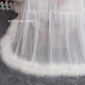 May include: A white, sheer garment with a fluffy, feathered trim. The garment has multiple layers and a ruffled design. The text "FashionGoodieUS" is visible on the left side of the image.