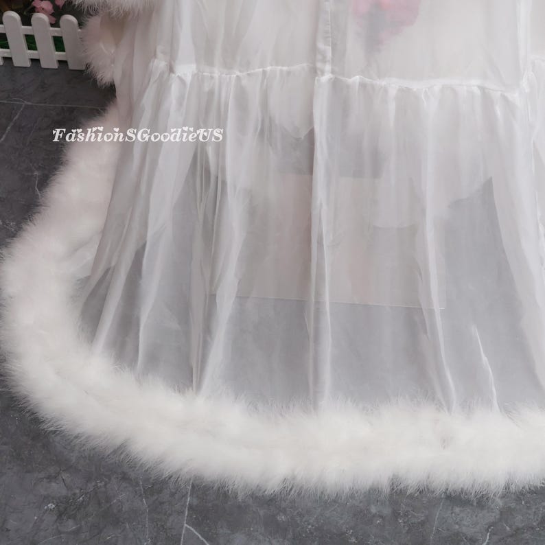 May include: A white, sheer garment with a fluffy, feathered trim. The garment has multiple layers and a ruffled design. The text "FashionGoodieUS" is visible on the left side of the image.