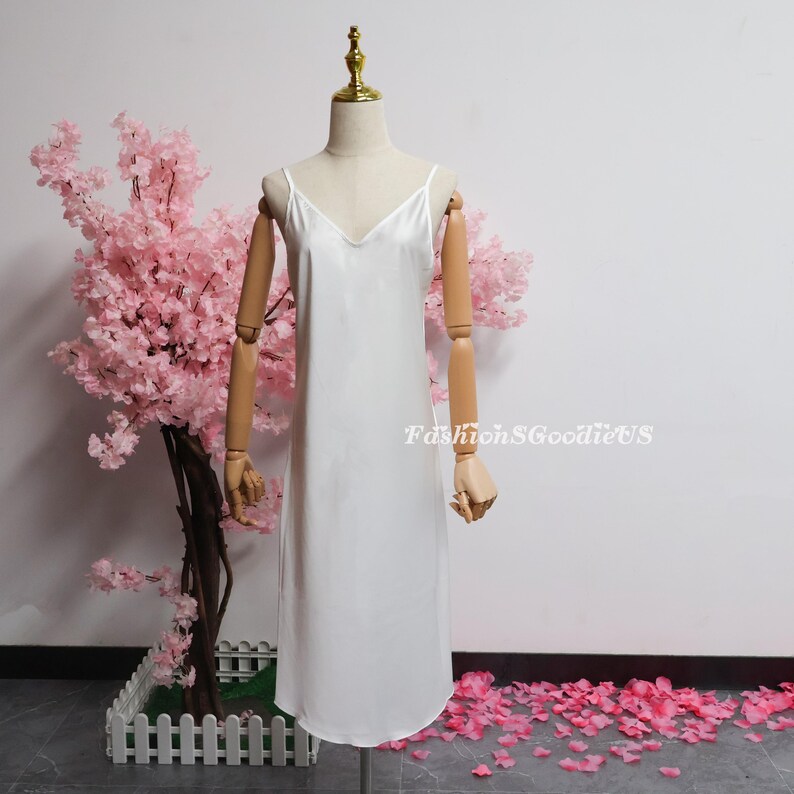 May include: A white satin slip dress with a V-neck and spaghetti straps is displayed on a mannequin. The dress is knee-length and has a slight A-line shape. The text "FashionSGoodieUS" is visible on the right side of the dress.