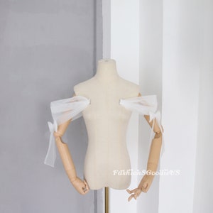 May include: White tulle off-the-shoulder sleeves with bows on the ends. The sleeves are on a white dress form.