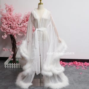 May include: A sheer white robe with a V-neck and a matching belt. The robe has long sleeves and is trimmed with fluffy white feathers along the hem and cuffs. The robe is displayed on a mannequin. The background includes pink flowers.