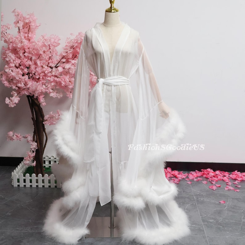 May include: A sheer white robe with a V-neck and a matching belt. The robe has long sleeves and is trimmed with fluffy white feathers along the hem and cuffs. The robe is displayed on a mannequin. The background includes pink flowers.
