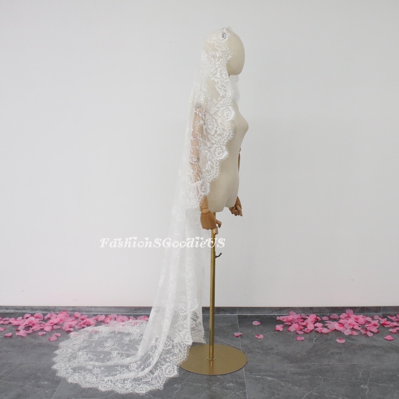 May include: A white lace wedding veil with a scalloped edge. The veil is draped over a mannequin and is displayed on a gold stand. Pink flower petals are scattered on the floor.