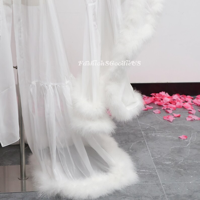 May include: A white, sheer robe with feather trim. The robe is made of a lightweight fabric and has a delicate design. The text "FashionSGoodieUS" is visible on the robe.