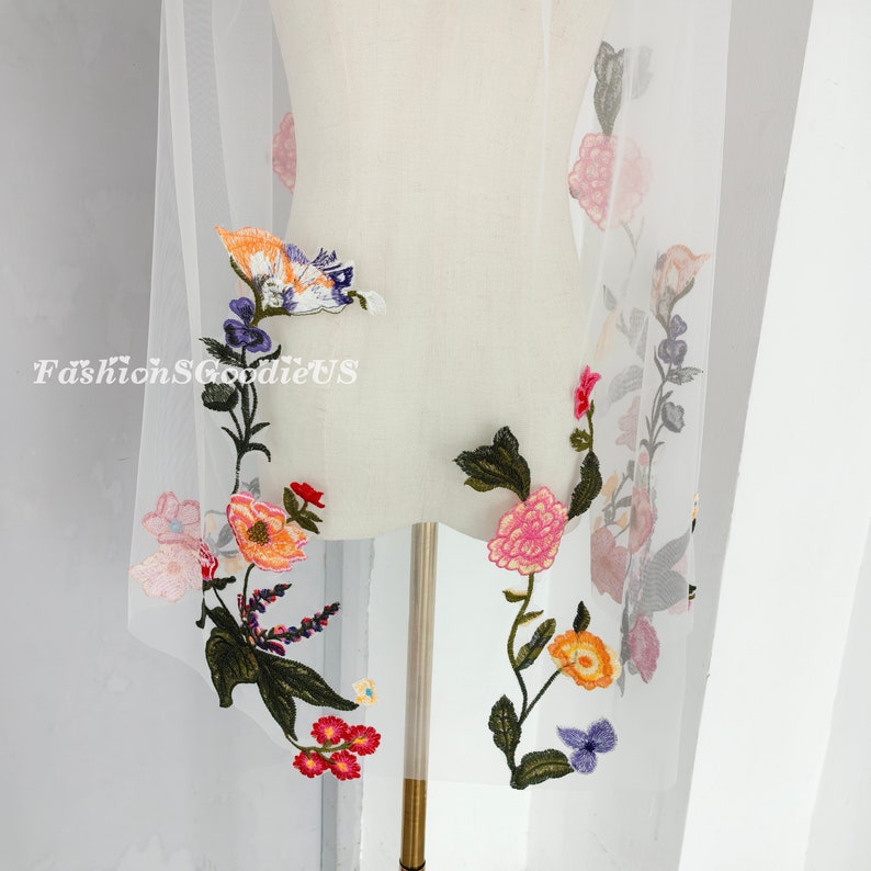 May include: White tulle fabric with colourful embroidered flowers. The flowers are in shades of pink, orange, yellow, blue, and green. The fabric is sheer and delicate.
