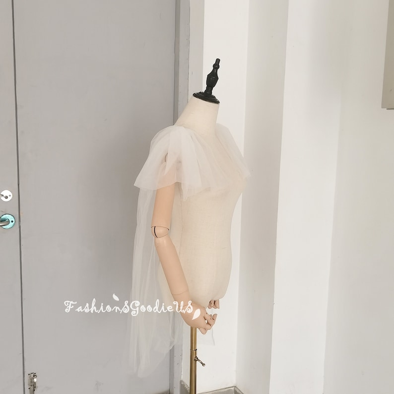 May include: A white tulle bridal cape with a ruffled neckline and short sleeves. The cape is draped over a white dress form.