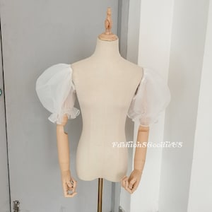 May include: White, sheer puff sleeves with ruffled cuffs displayed on a dress form. The sleeves are made of a lightweight, airy fabric. The dress form is beige and has wooden arms and a wooden neck.