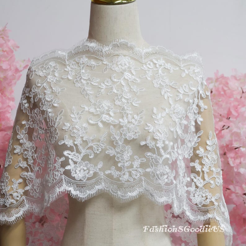 May include: A white lace fabric featuring an intricate floral embroidery pattern with a scalloped edge. The delicate mesh material is draped over a mannequin, showcasing the detailed stitching and the text FashionSGoodieUS in the corner.