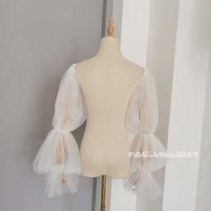 May include: White tulle off-the-shoulder sleeves with puffed and ruffled details. The sleeves are long and flowy, with a delicate and romantic look.