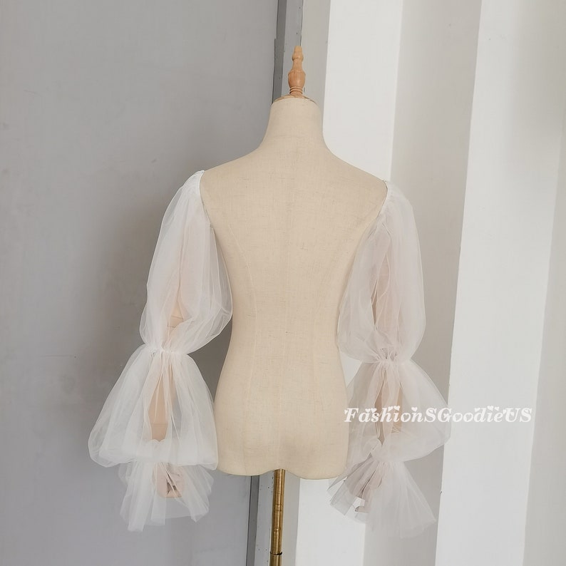 May include: White tulle off-the-shoulder sleeves with puffed and ruffled details. The sleeves are long and flowy, with a delicate and romantic look.