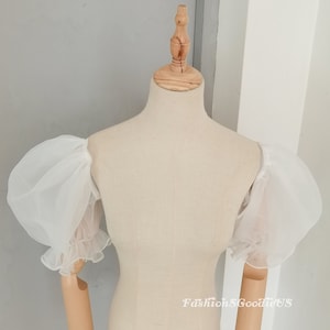 May include: White, sheer, puff sleeves with ruffled cuffs. The sleeves are displayed on a dress form. The text "FashionSGoodieUS" is visible in the lower right corner. The sleeves are made of a lightweight, airy fabric.