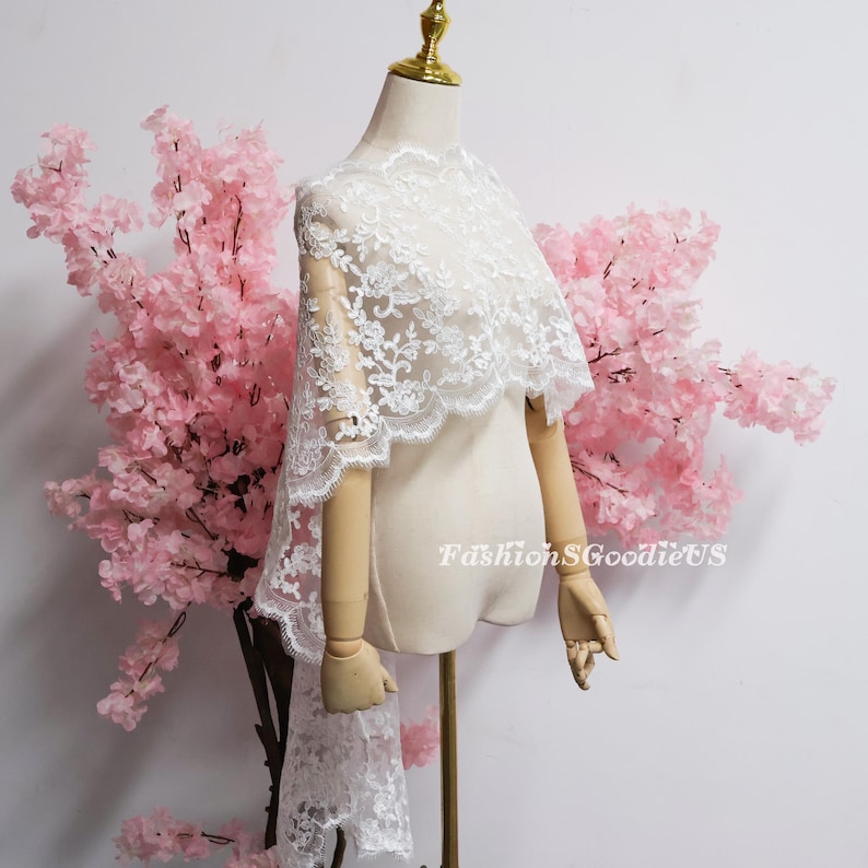 May include: A white lace bridal capelet featuring intricate floral embroidery and a scalloped edge, draped over a mannequin. The fabric is sheer with detailed needlework patterns throughout. The text FashionSGoodieUS is visible on the right side.