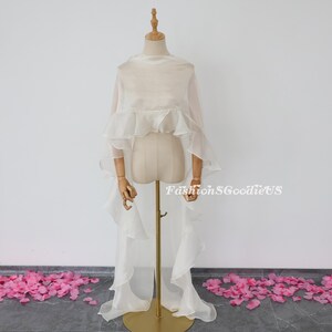 Pu&ograve; includere: Un scialle bianco di chiffon con volant e una lunga coda. Lo scialle &egrave; drappeggiato su un manichino bianco. Il manichino &egrave; in piedi su un supporto dorato. Ci sono petali di fiori rosa sparsi sul pavimento.