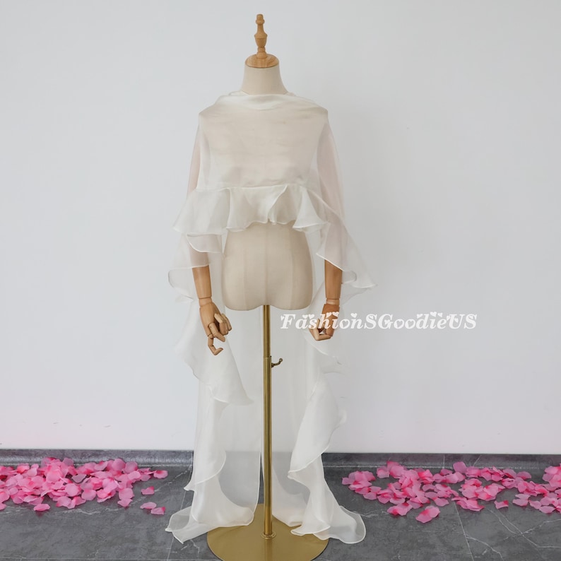 Pu&ograve; includere: Un scialle bianco di chiffon con volant e una lunga coda. Lo scialle &egrave; drappeggiato su un manichino bianco. Il manichino &egrave; in piedi su un supporto dorato. Ci sono petali di fiori rosa sparsi sul pavimento.