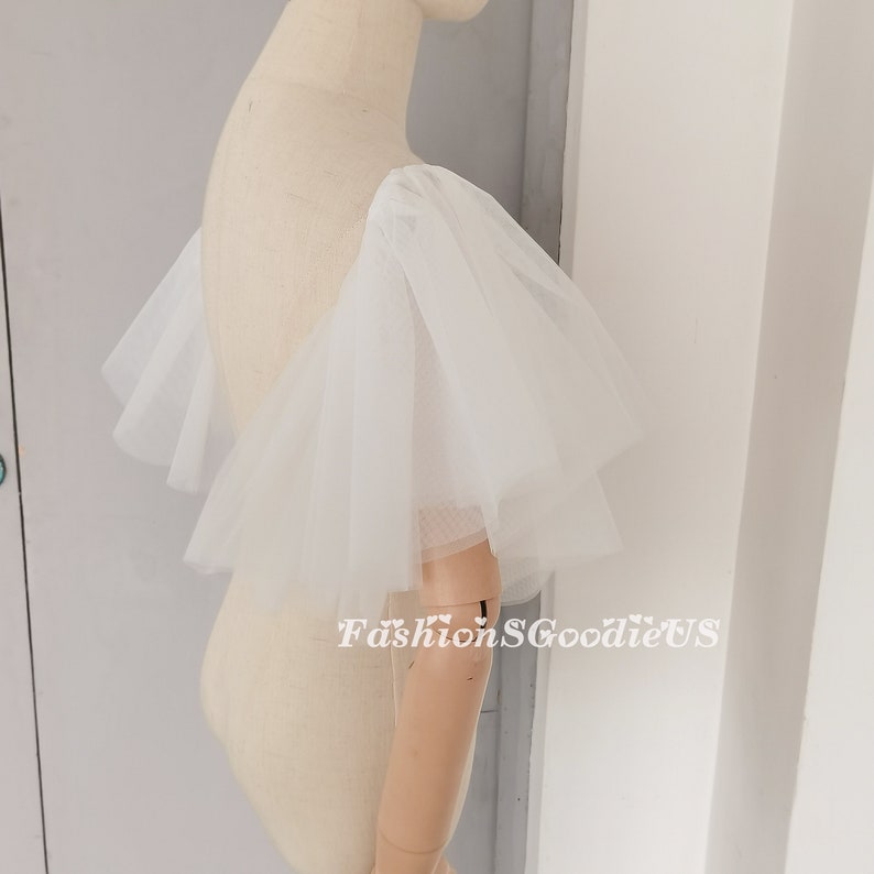 Pu&ograve; includere: Maniche in tulle bianco con un design a balze. Le maniche sono attaccate a un manichino bianco.