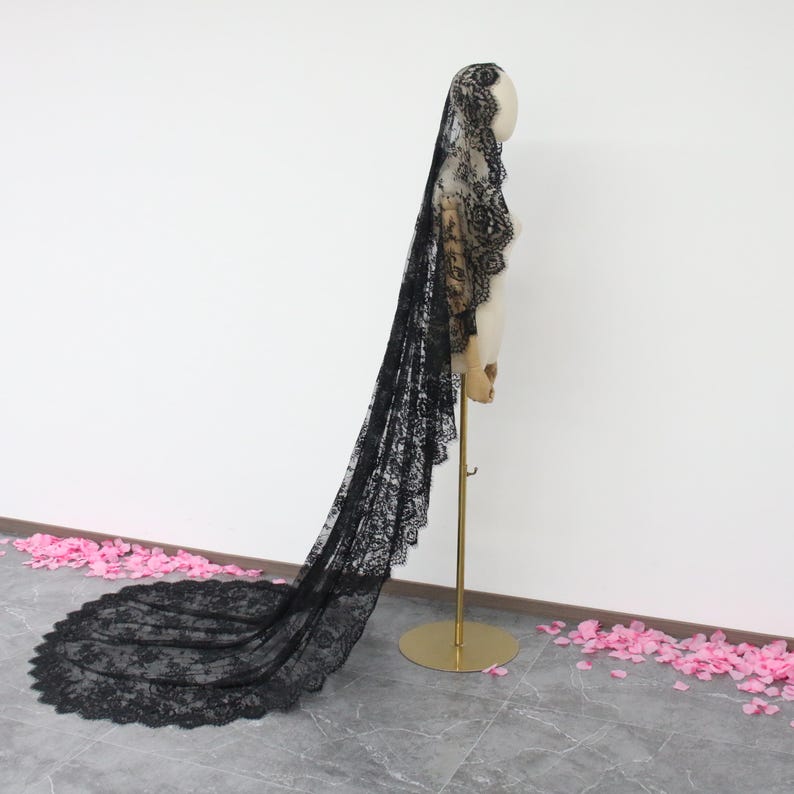 May include: A long black lace veil with a scalloped edge. The veil is draped over a mannequin and is displayed against a white background.