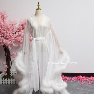 May include: A white, sheer robe with a tie belt and fluffy feather trim. The robe is displayed on a mannequin, with a pink floral arrangement in the background. The text "FashionSGoodieUS" is visible on the robe.