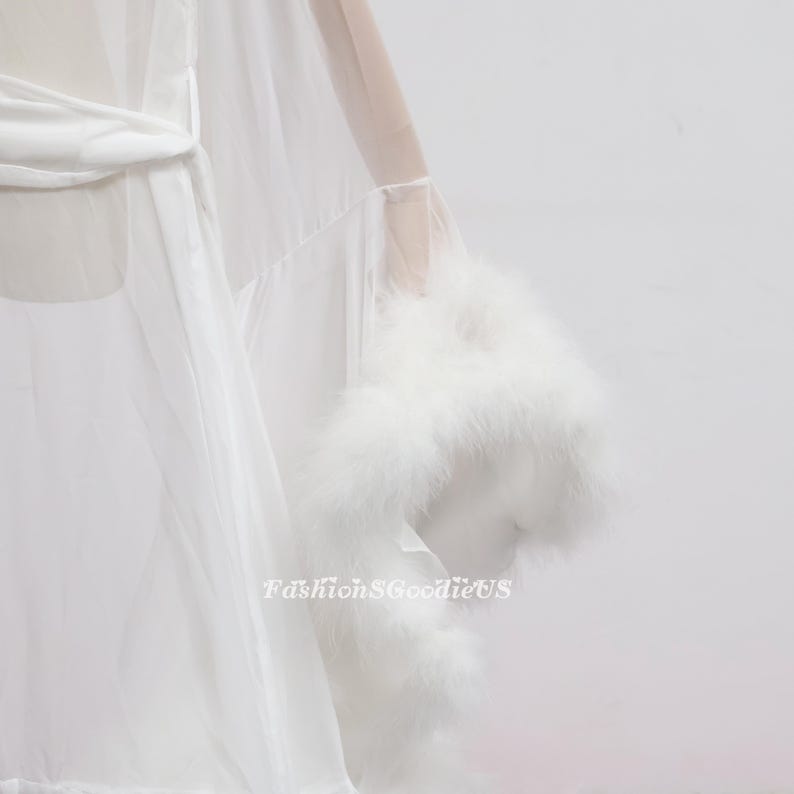 May include: White sheer robe with a satin belt and feather trim on the sleeves. The text "FashionGoodieUS" is visible on the lower right side of the image. The robe is made of a lightweight fabric.