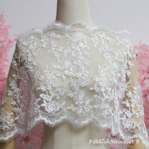 May include: A white lace fabric featuring an intricate floral embroidery pattern with scalloped edges. The delicate mesh material is draped over a mannequin, showcasing the detailed stitching and the text FashionSgoodieUS in the bottom corner.