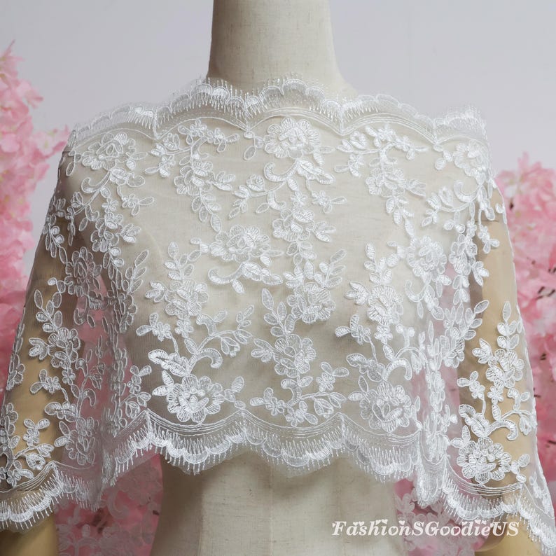 May include: A white lace fabric featuring an intricate floral embroidery pattern with scalloped edges. The delicate mesh material is draped over a mannequin, showcasing the detailed stitching and the text FashionSgoodieUS in the bottom corner.