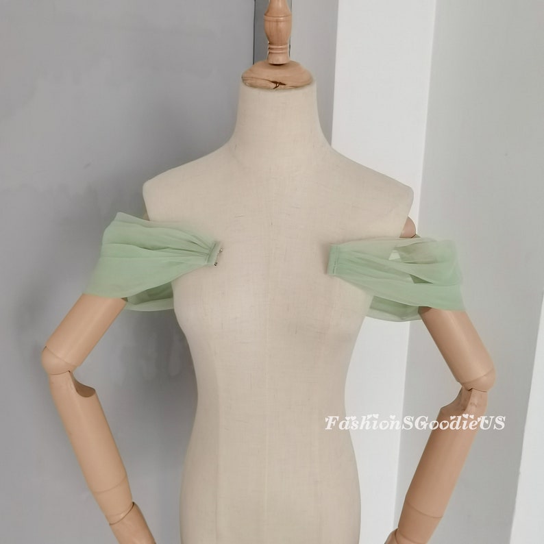 May include: A pair of off-the-shoulder tulle sleeves in a light green color. The sleeves are gathered at the shoulders and have a delicate, flowing look.  FashionSGoodieUS is printed on the bottom right corner of the image.