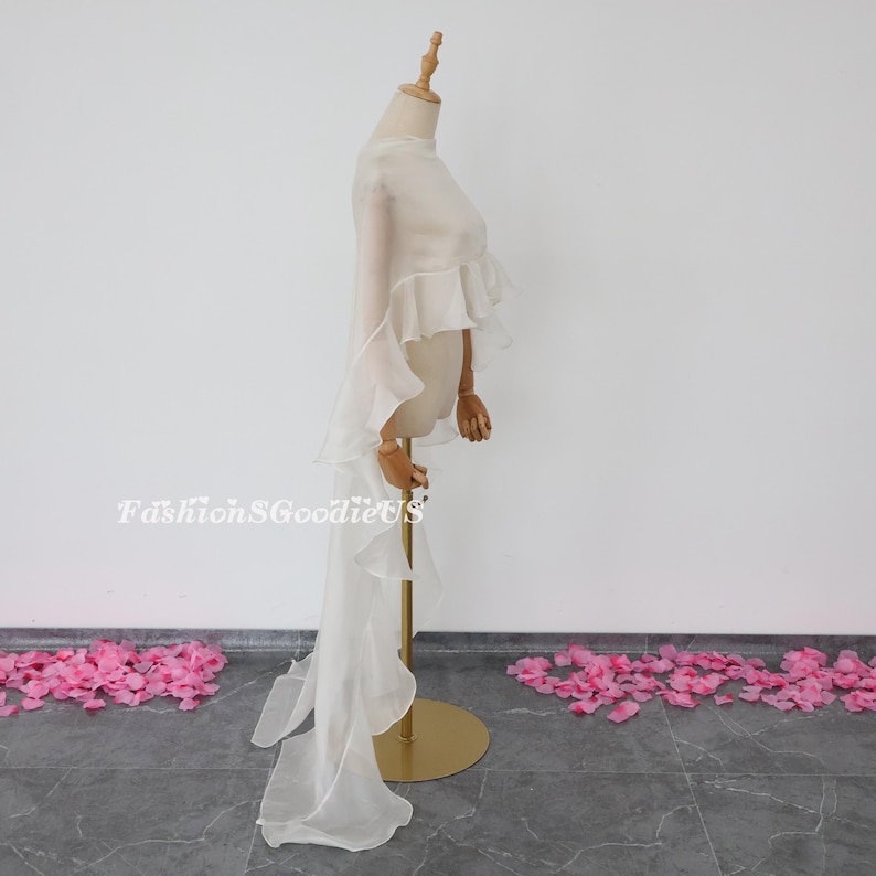 Pu&ograve; includere: Una cappa bianca in chiffon con bordi a balze e una lunga coda. La cappa &egrave; drappeggiata su un manichino ed &egrave; esposta su uno sfondo bianco.