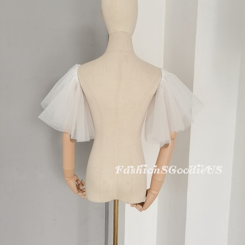 Pu&ograve; includere: Manichini con maniche in tulle bianco con un design a balze. Le maniche sono mostrate su un manichino bianco.