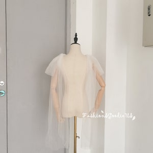 May include: A white tulle cape with ruffled shoulders. The cape is on a dress form.