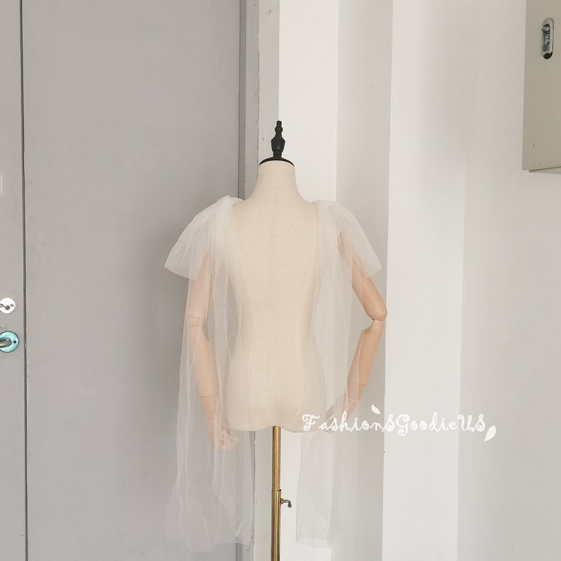 May include: A white tulle cape with ruffled shoulders. The cape is on a dress form.