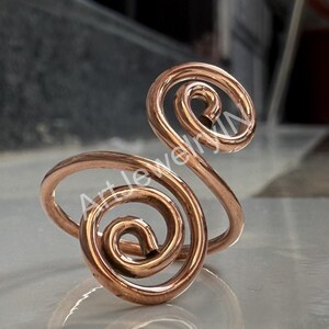 Op de afbeelding: Een koperkleurige ring met een uniek spiraalontwerp. De ring heeft twee prominente, open spiralen, de ene groter dan de andere, verbonden door een band. Het metaal heeft een gepolijste afwerking, die licht reflecteert.