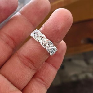 May include: A silver ring featuring a detailed braided design that mimics the appearance of woven rope. The band has a polished metallic finish with intricate twisted strands that create a textured, continuous pattern around the entire circumference.