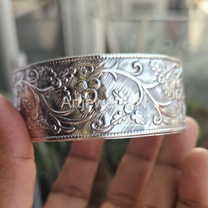 Op de afbeelding: Een brede, zilverkleurige manchetarmband met een ingewikkeld bloemenontwerp. De armband heeft gedetailleerde gravures van bloemen en bladeren, waardoor een textuur ontstaat. Het metaal heeft een gepolijste afwerking, die licht reflecteert.