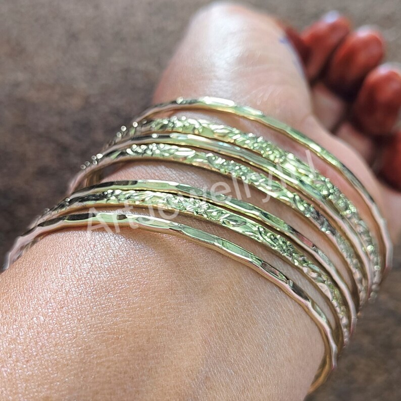 May include: A set of eight gold-toned bangles with an etched floral design. The bangles are stacked on a person's wrist.