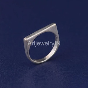 Può includere: Un anello d'argento con una fascia rettangolare. La fascia è piatta e ha un bordo leggermente arrotondato. L'anello è semplice e di design moderno.