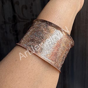 May include: Wide copper cuff bracelet with an intricate, swirling pattern. The cuff is a warm, metallic color and wraps around the wrist. The design is detailed and appears to be etched or embossed into the metal.