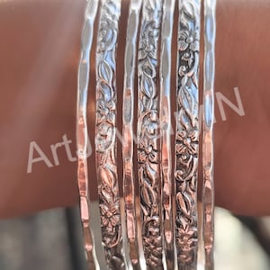 Puede incluir: Conjunto de siete pulseras de plata con diseños florales. Las pulseras están apiladas una encima de la otra y tienen una textura martillada.