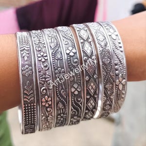 Pode incluir: Um conjunto de pulseiras de prata com intrincados desenhos florais e geométricos. As pulseiras estão empilhadas no pulso de uma pessoa.