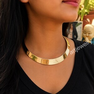 Puede incluir: Un collar tipo gargantilla de metal en tono dorado pulido con un diseño de banda ancha, curva y lisa que se ajusta al cuello. La pieza presenta un acabado brillante y reflectante, con una estructura sólida y continua sin cierres visibles.