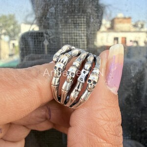 Puede incluir: Un anillo plateado con forma de mano de esqueleto, con una calavera en cada dedo. El anillo es detallado y parece estar hecho de metal. El anillo se lleva en un dedo.