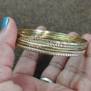 Pode incluir: Um conjunto de pulseiras douradas com vários desenhos gravados. As pulseiras são seguradas em uma mão com esmalte de unha azul. As pulseiras têm diferentes texturas, incluindo superfícies pontilhadas, trançadas e estampadas. As joias são exibidas em um fundo marrom.