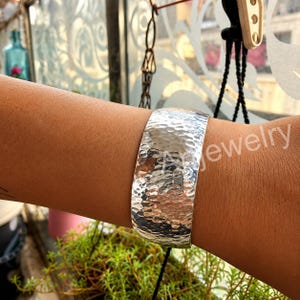 Pode incluir: Uma pulseira larga de tom prateado com uma superfície martelada e texturizada que cria uma aparência reflexiva e irregular. A banda de metal tem uma estrutura curva e sólida que envolve o pulso, exibindo um acabamento polido.