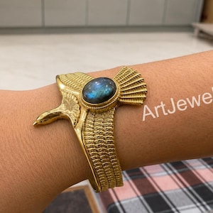 May include: A gold-toned metal cuff bracelet shaped like a bird with outstretched wings, featuring a central oval-shaped iridescent blue labradorite gemstone. The surface of the metal is textured with fine lines to resemble feathers. Text reads ArtJewelry.