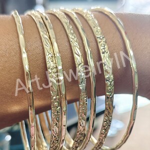 May include: A set of eight gold-toned bangles with floral designs. The bangles are stacked on a person's arm.