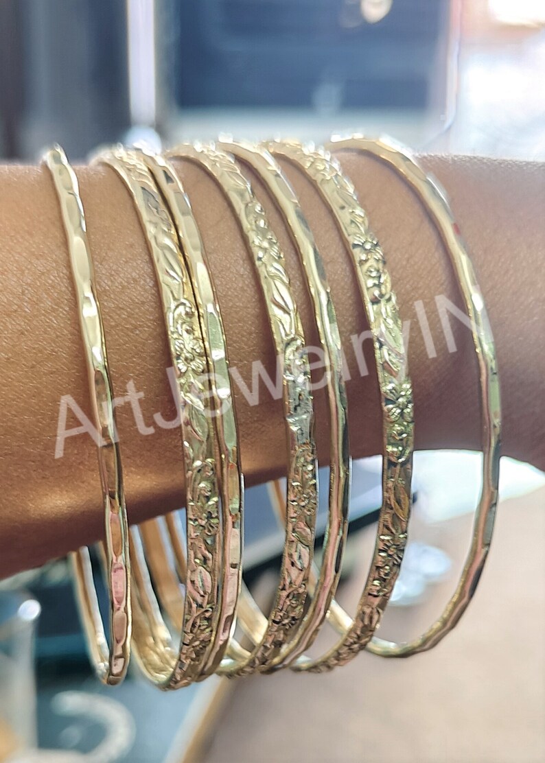 May include: A set of eight gold-toned bangles with floral designs. The bangles are stacked on a person's arm.