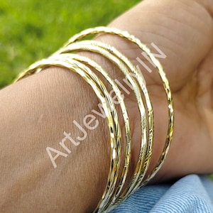 Puede incluir: Un conjunto de seis pulseras doradas con un patrón texturizado en forma de diamante. Las pulseras están apiladas en la muñeca de una persona.