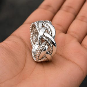 Può includere: Un anello in argento lucido caratterizzato da un design complesso e intrecciato che combina fasce lisce e piatte con texture ritorte simili a una corda. Il metallo presenta una finitura brillante e riflettente, creando un aspetto multistrato con profondità visibile.